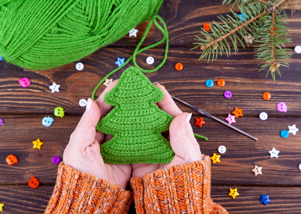 A pair of hands holding a small green crochet Christmas tree above a wooden table with multicolored stars and a green yarn ball on.