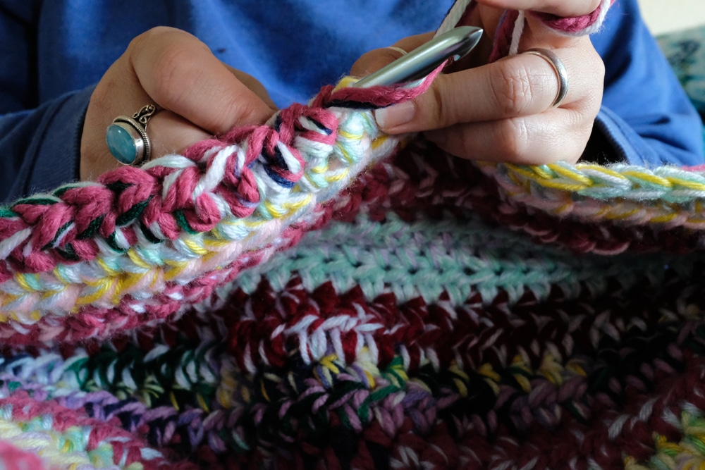 A woman’s hands with knitting needles adding to a multi-colored crochet blanket featuring a range of stitch designs. 
