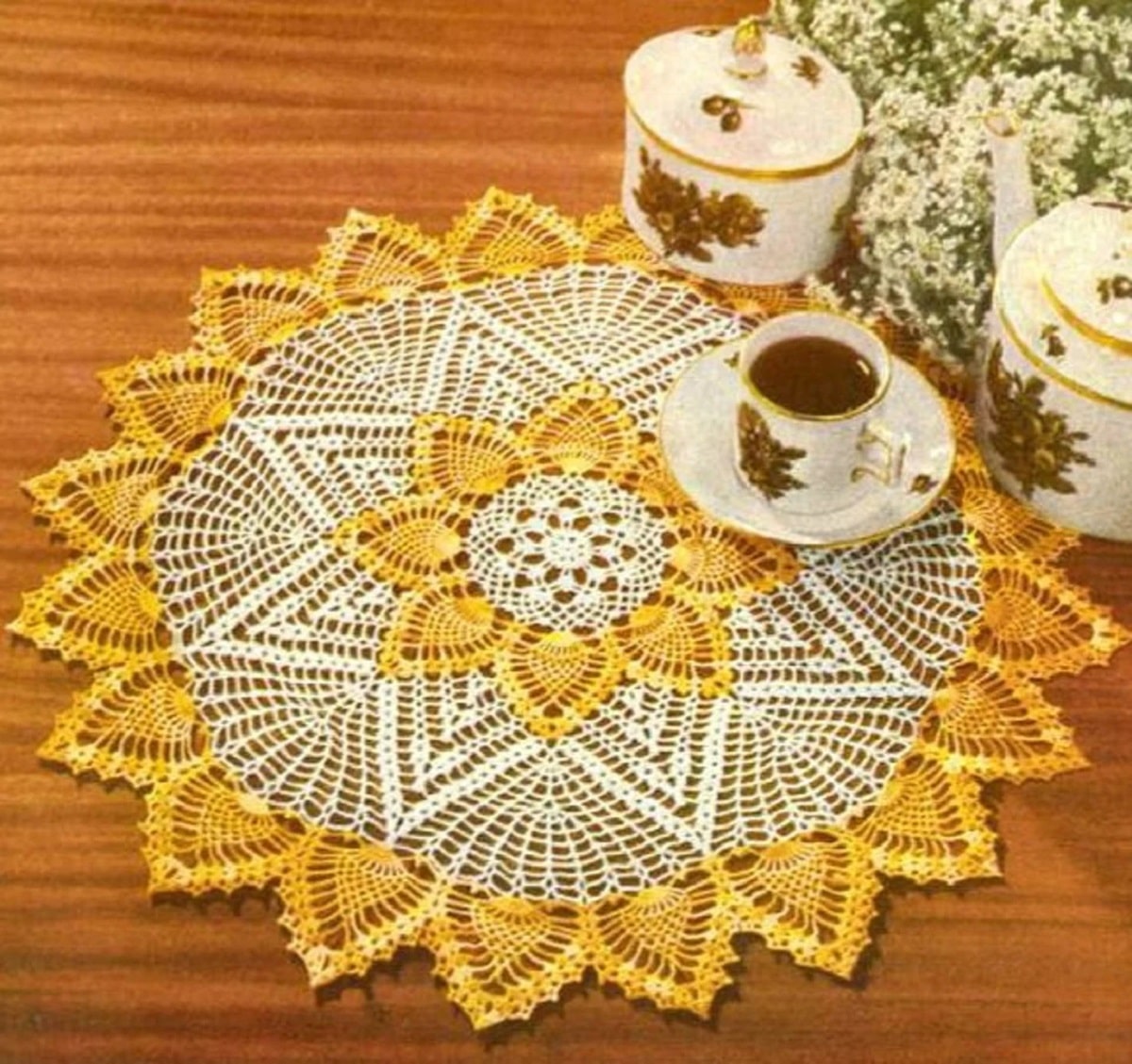  Yellow pineapples in petals in the center of this cream crochet doily, with yellow petals around the entire circle on a wooden table.