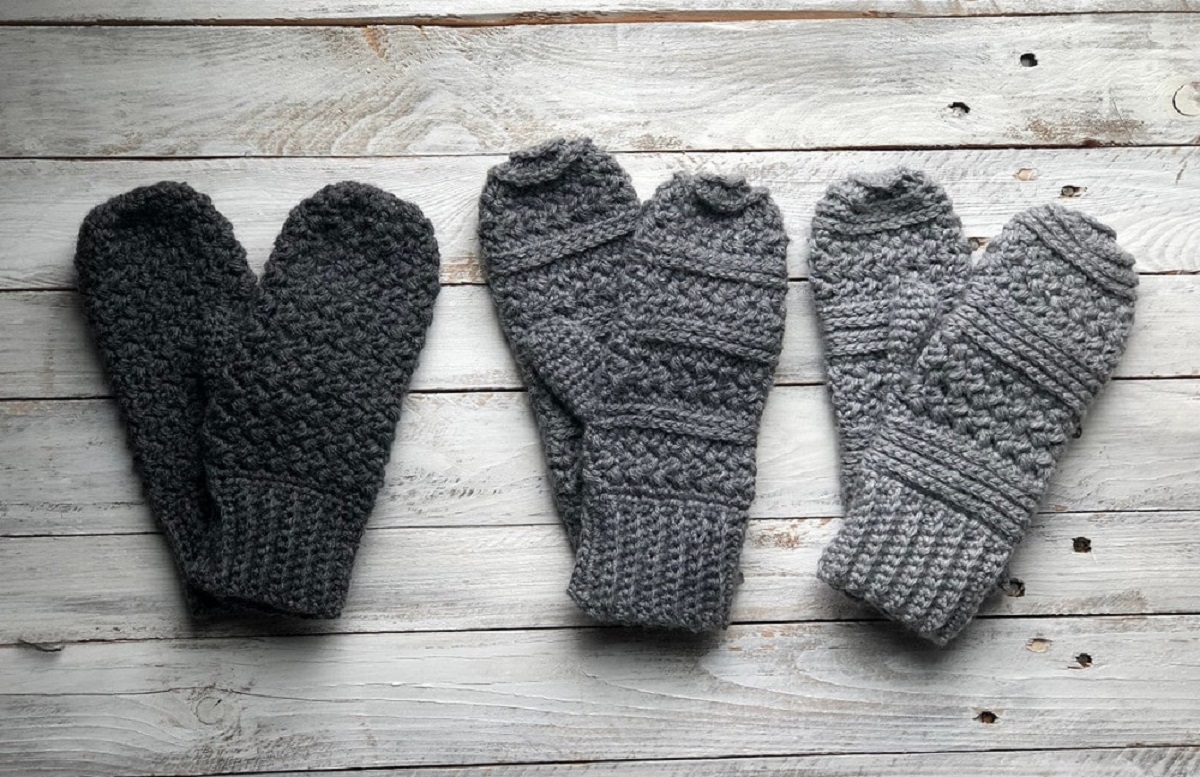 Black, dark gray, and light gray mittens with vertical lines and cable stitching on a pale wooden background.