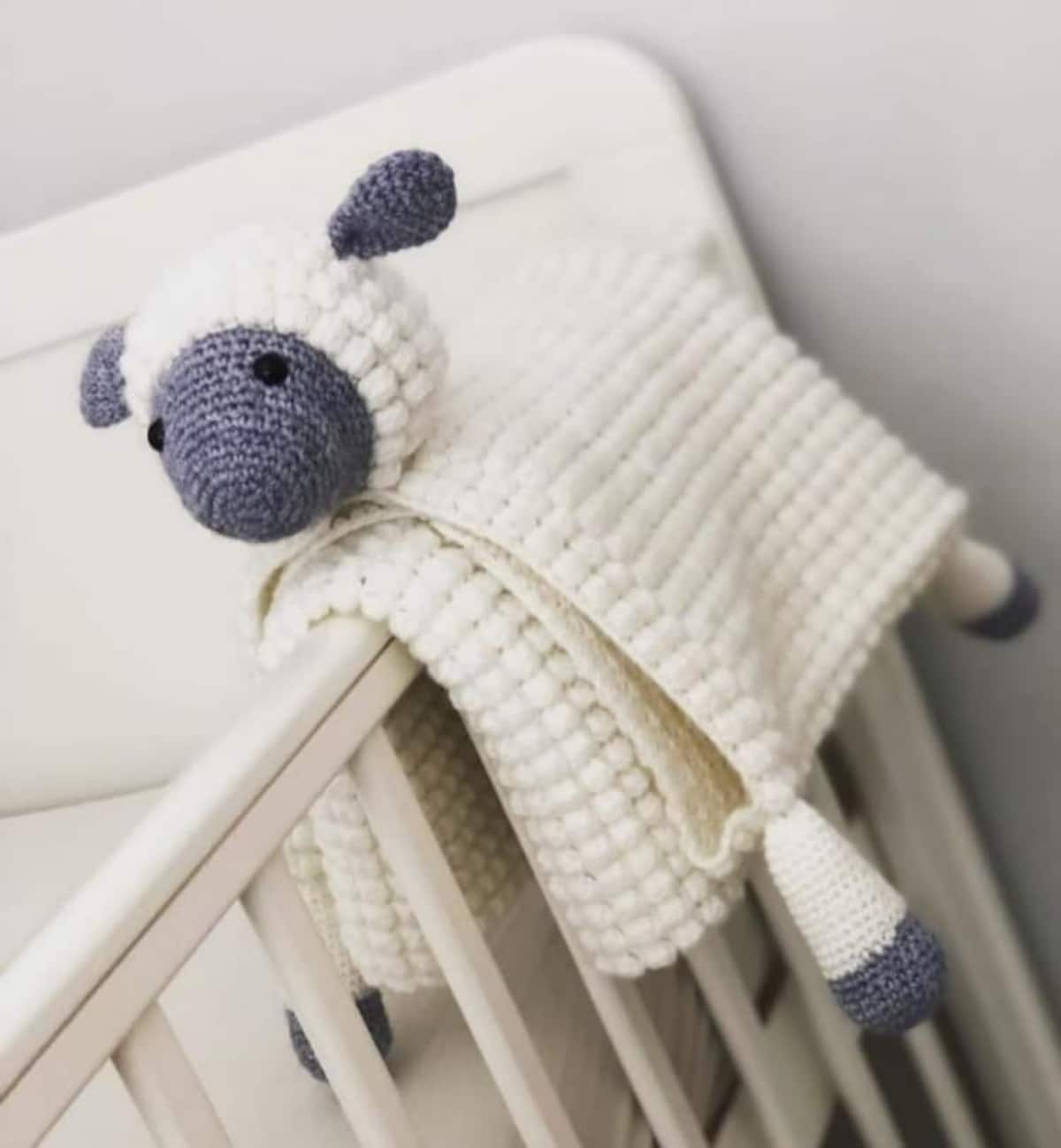 White crochet textured blanket with a stuffed gray and white lamb’s face and legs attached folded over a crib’s railings.