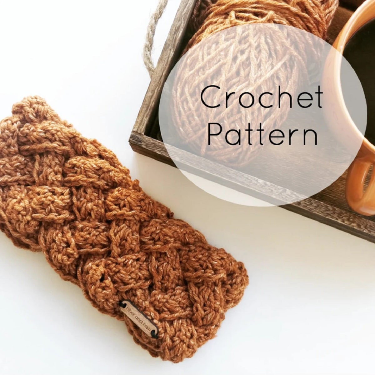 A tan colored crochet headband using a chunky braided design next to a wooden tray with yarn and a cup of coffee on.