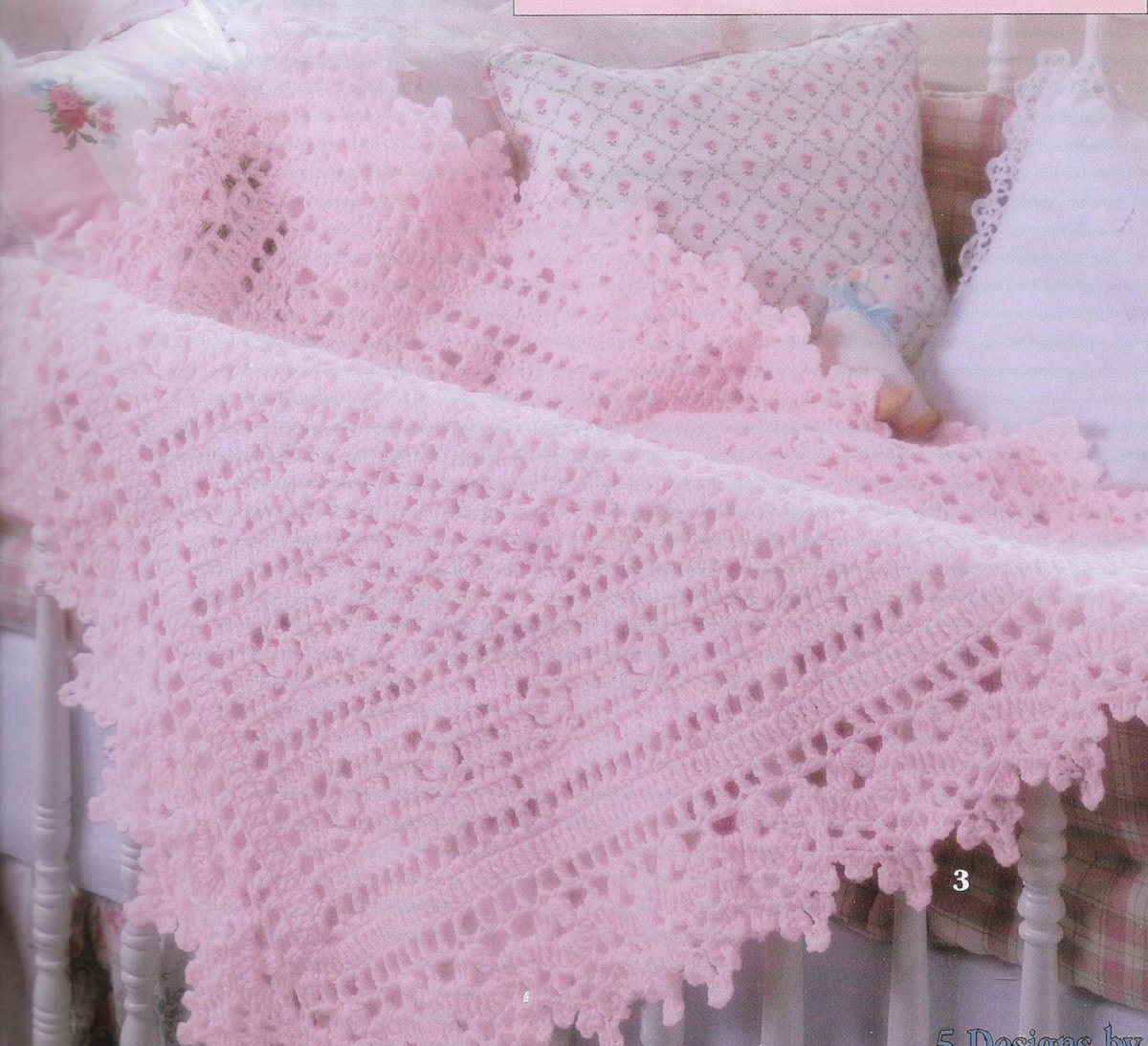 Baby pink crochet blanket with a lace style trim around the side hanging over a cribs railings in a diamond shape.