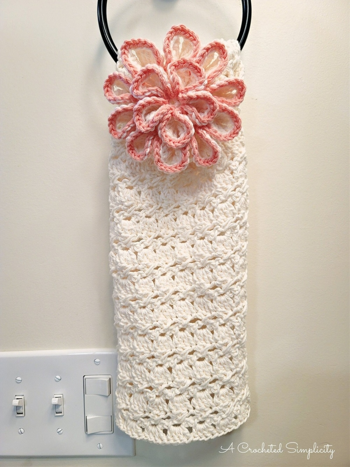 A white crochet hand towel with a large 3-D pink flower at the top folded over a towel hoop.