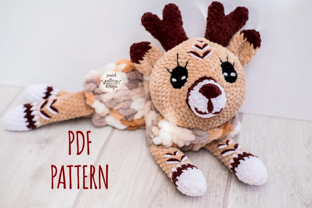 A brown crochet stuffed deer with dark brown antlers, and a multicolored back, lying with its legs spread out.