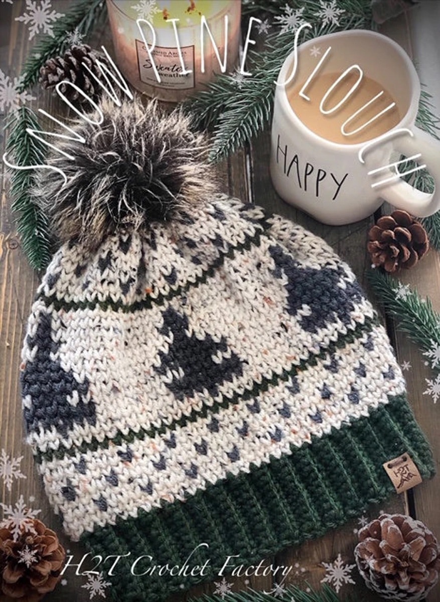 White and green alpine style slouchy beanie with christmas trees and small green hearts stitched in and a large gray fluffy bobble on top.