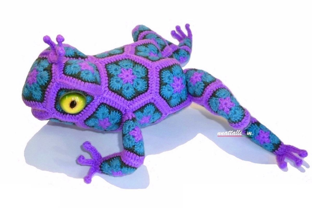 Crochet frog laying down with its legs spread on a white background. Purple frog features blue and purple hexagon flowers stitched over its entire body.