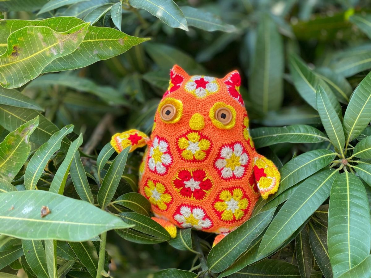 Bright orange crochet owl with yellow and brown eyes and African flowers stitched across its body and head sitting in some leaves.
