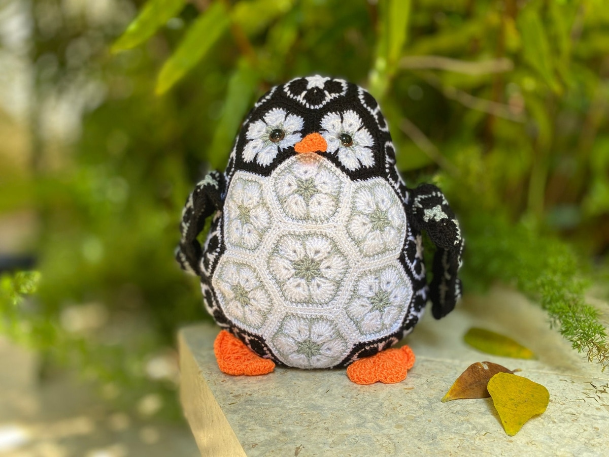 Crochet black and white penguin with African flower pattern over its body with flowers for eyes and an orange nose and feet standing by some green leaves.