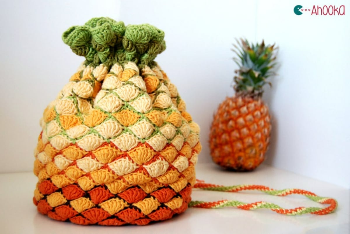 An orange, red, and yellow pineapple style crochet bag with a green banding on top and an orange and green strap behind it.