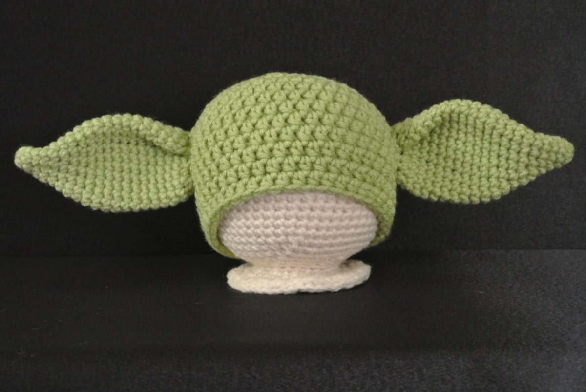 A crochet mannequin wearing a green beanie hat with large Yoda ears either side on a black background.