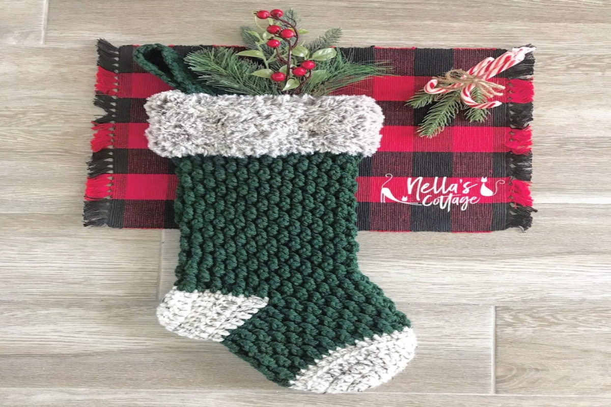 Green alpine-style stocking with white banding laying on a pale wooden floor with a red and black blanket and Christmas decorations. 