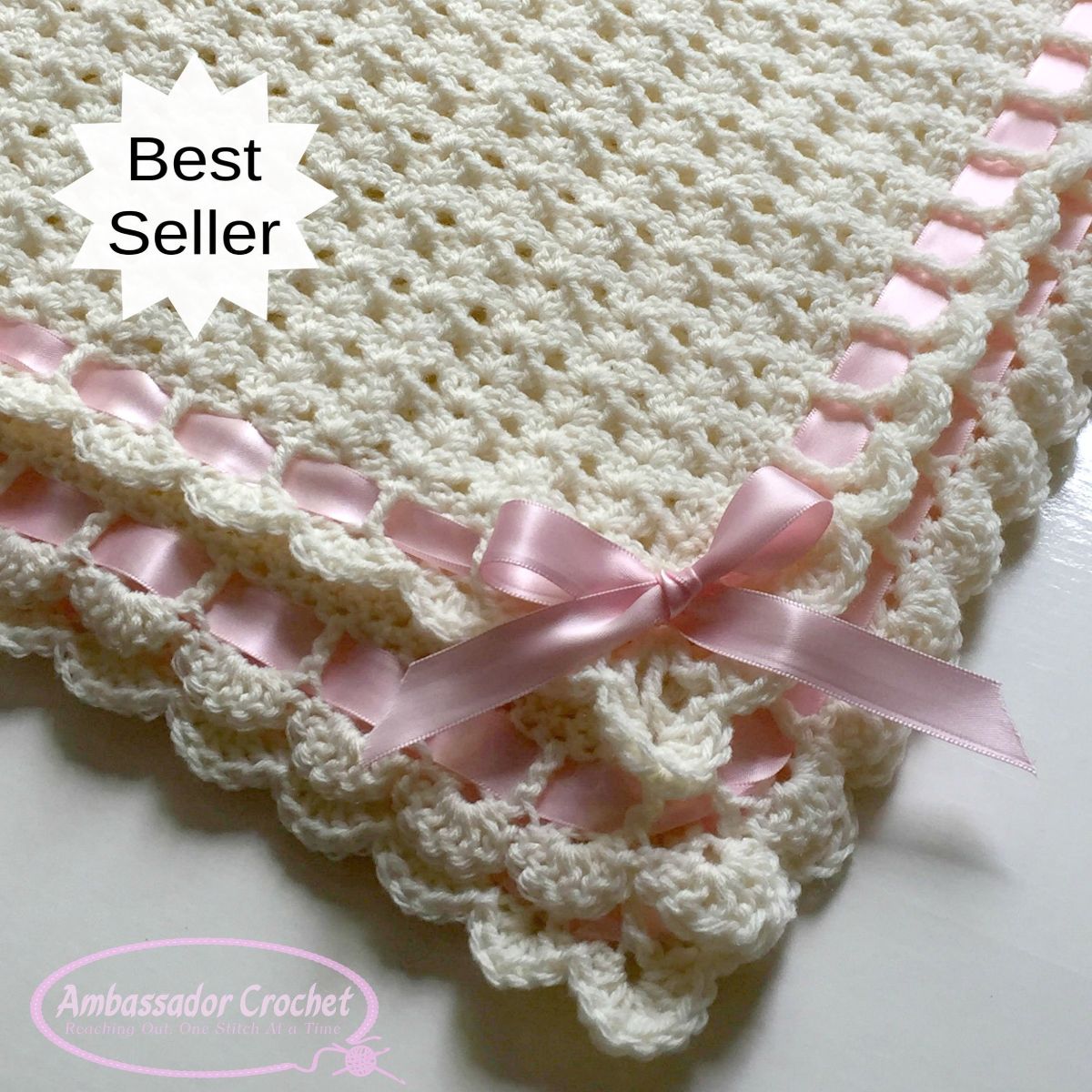 White crochet baby blanket with a pink satin ribbon stitched around the outside with a pink bow in the corner and a small lace trim underneath.