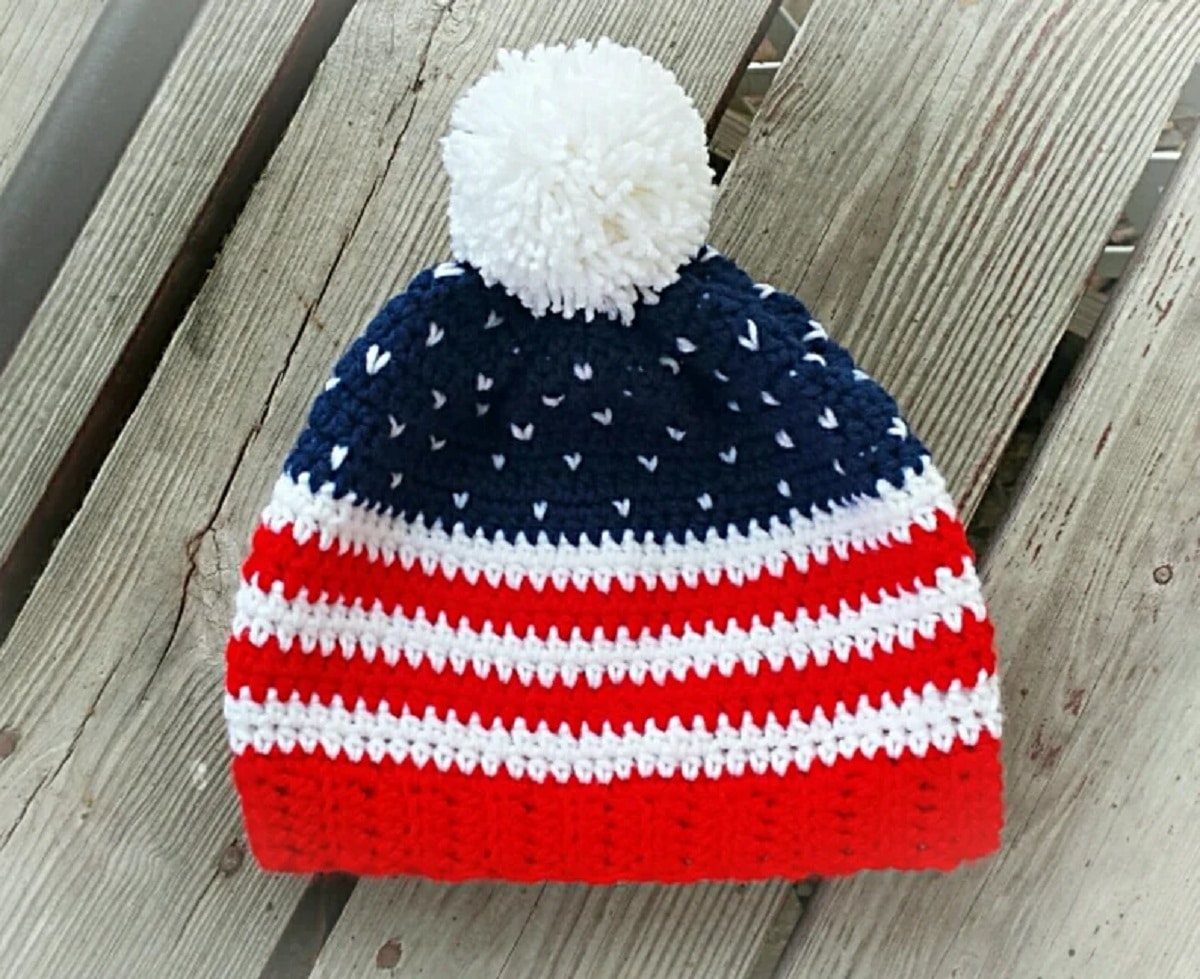 Crochet beanie hat with red and white stripes, a blue top with small white hearts, and white bobble on a wooden floor.