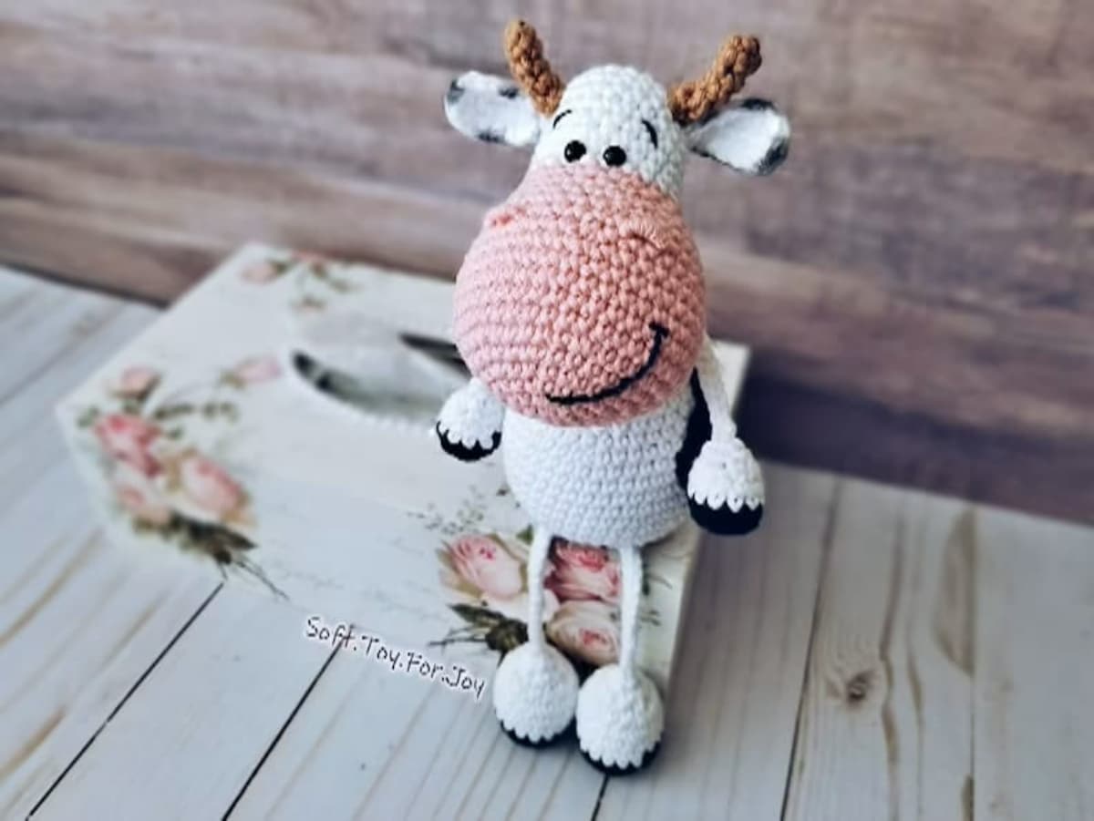  White crochet cow standing on two legs with an oversized pink nose, brown horns, and black detailing on its ears and feet.