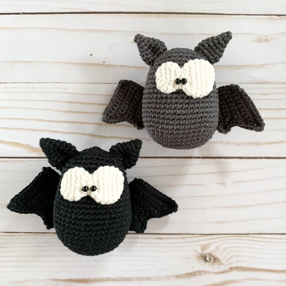 A black crochet bat with large white eyes next to a gray bat of the same style on a pale wooden background.