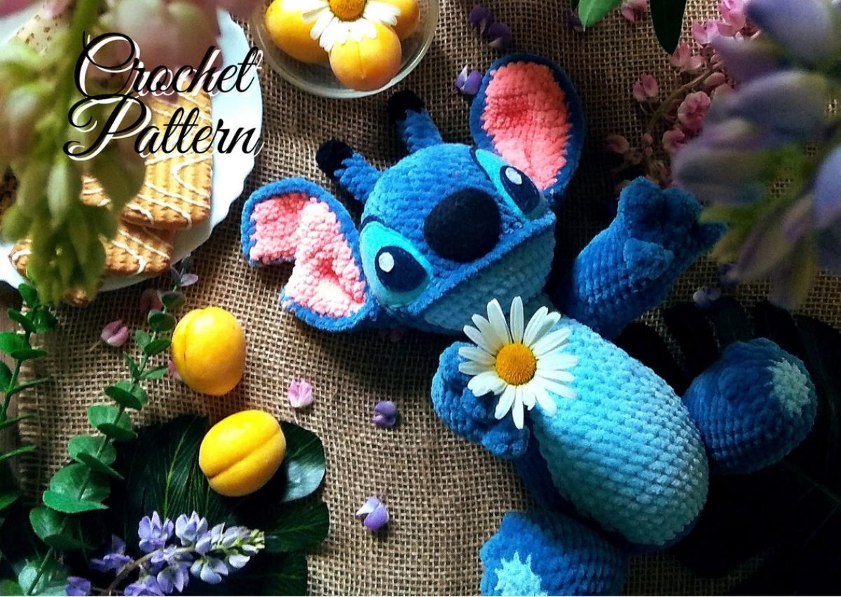 Large stuffed crochet Stitch pattern holding a daisy and lying on a brown background surrounded by fruit and foliage.