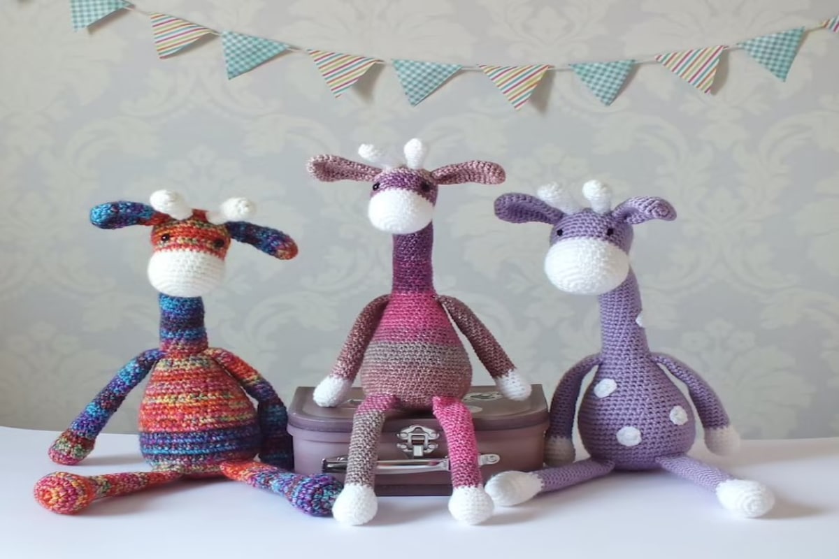 A blue and red crochet giraffe, purple and pink striped giraffe, and purple giraffe with white spots sitting next to each other with bunting behind them.