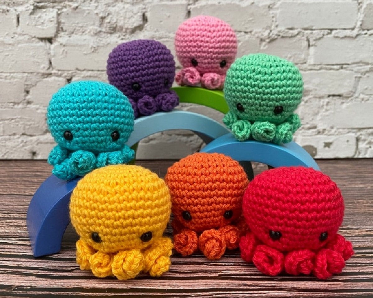 Seven crochet jellyfish in red, orange, yellow, blue, green, purple, and pink sitting on semicircles by a white wall.