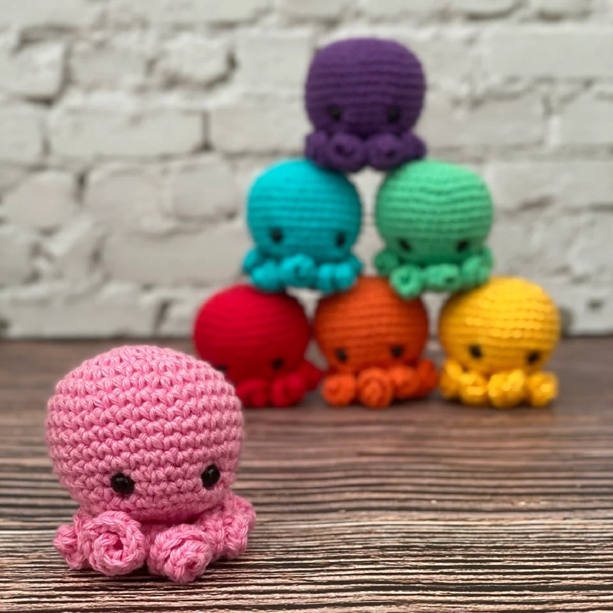  Pink crochet mini octopus with an oversized head and small tentacles standing in front of a pyramid of yellow, orange, red, blue, green, and purple octopuses. 