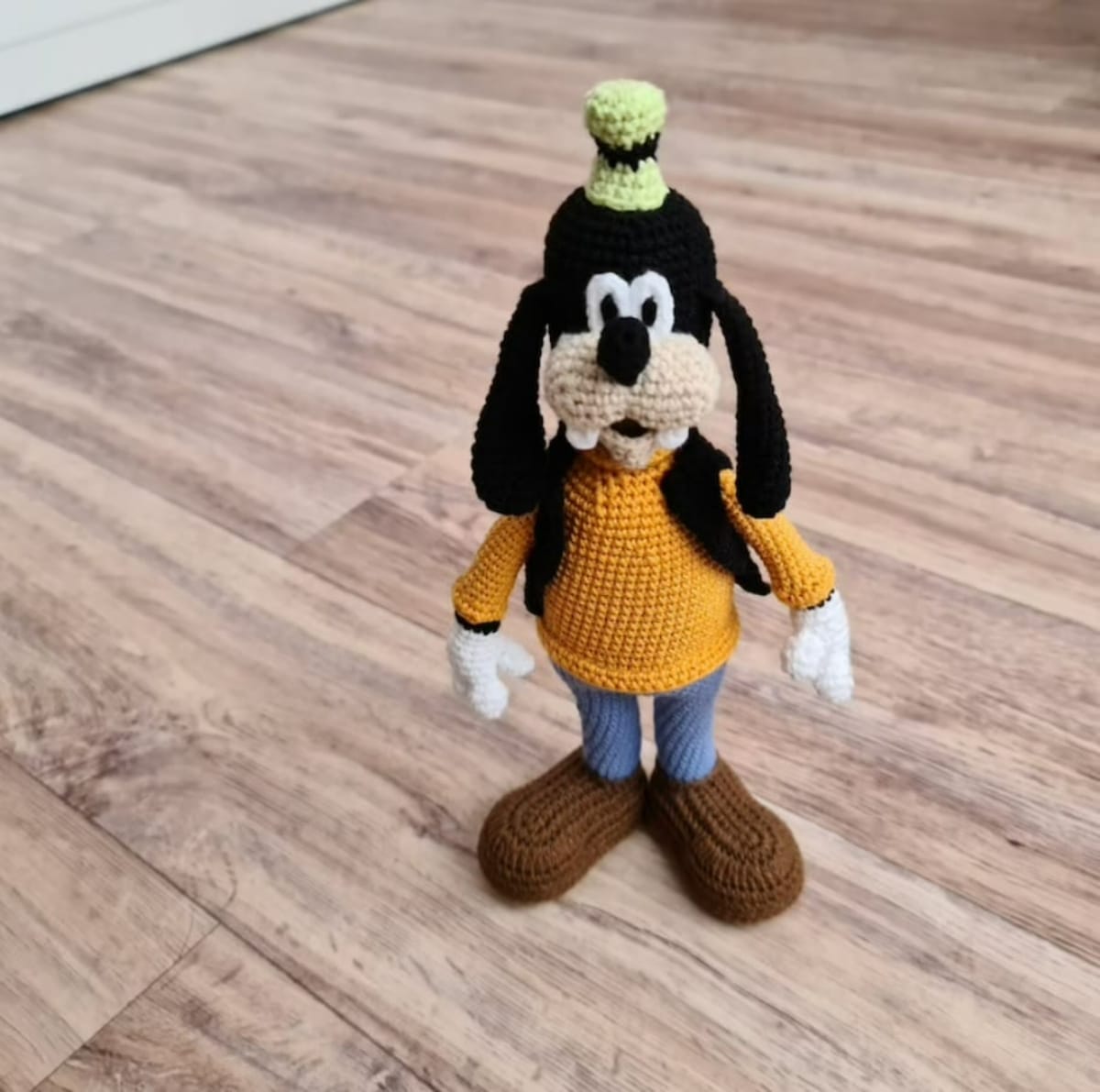 Stuffed crochet Goofy toy wearing a green hat, yellow jumper, blue trousers, and brown shoes standing on a wooden floor.