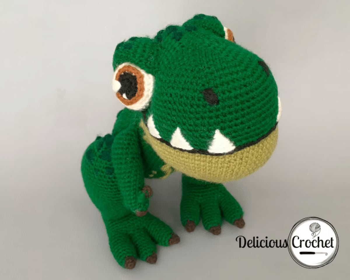 Dark green crochet stuffed T-Rex with an oversized head and large white and brown eyes sitting on a white background.
