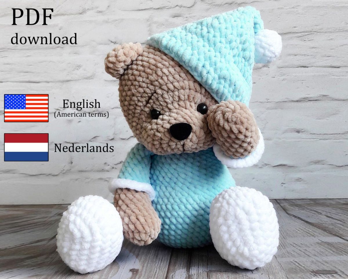 A brown crochet teddy bear wearing a blue night cap, night shirt, and white slippers sitting down on a gray wooden floor.