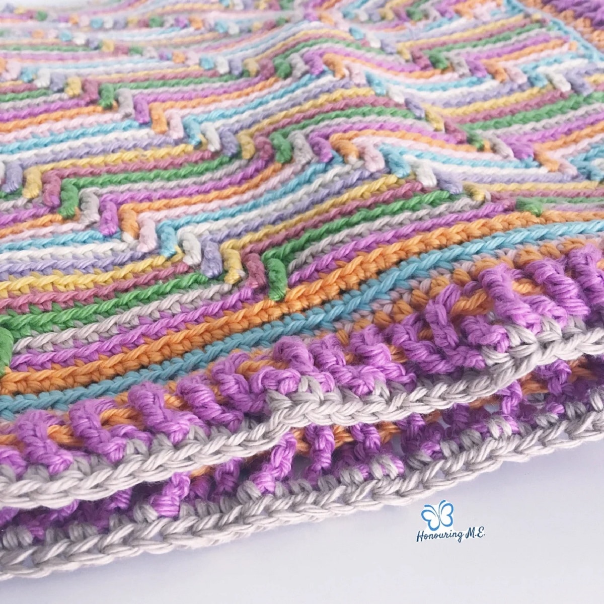  Pink, purple, green, orange, blue, and cream striped crochet blanket with a purple and cream trim on a white background.
