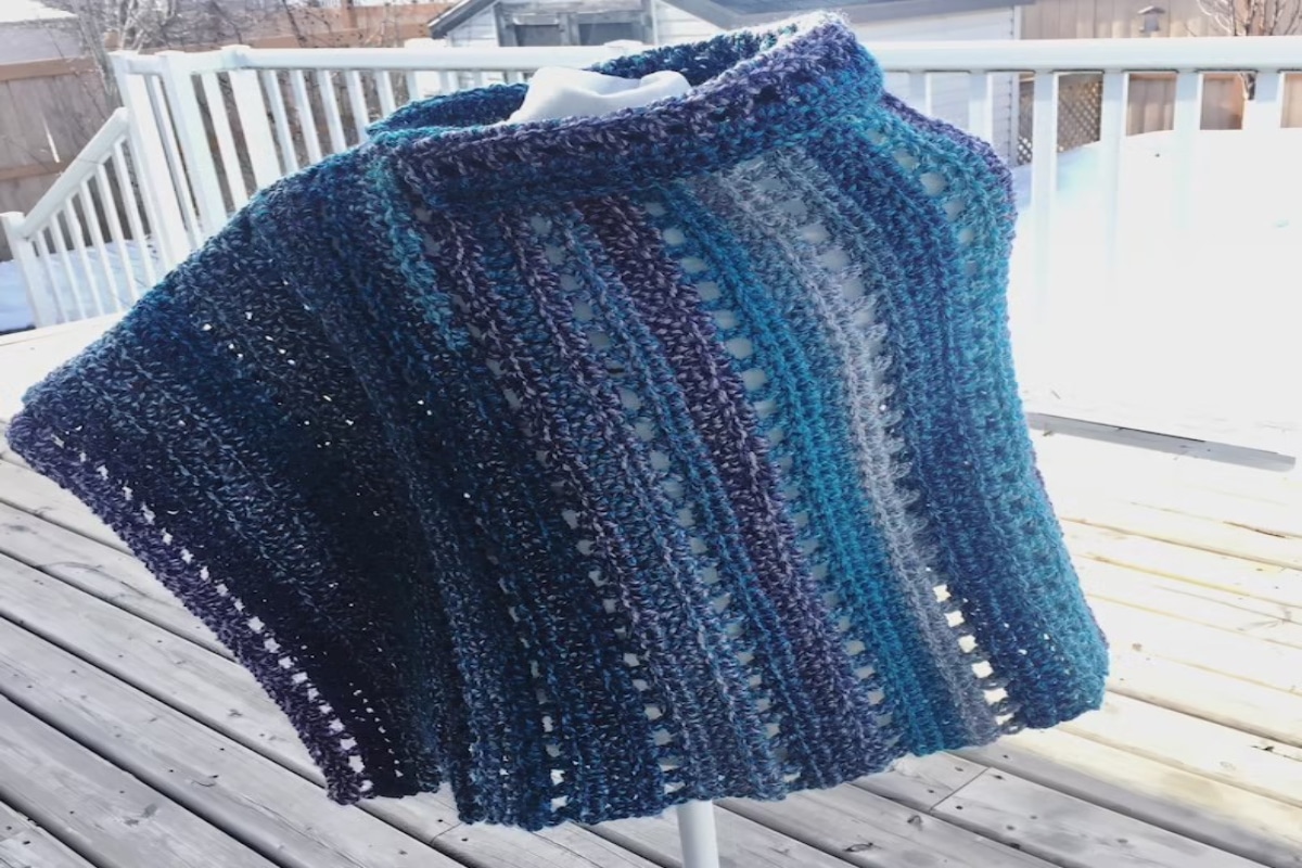 A vertical striped crochet poncho with various shades of blue and loose stitches in between each color and a high rounded neckline.