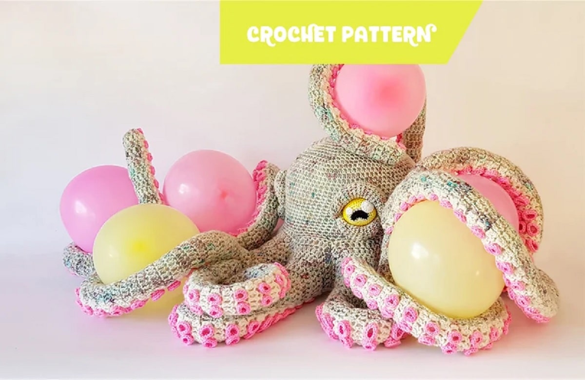 Large beige crochet octopus with cream tentacles with small pink bobbles on, holding yellow and pink balloons with its tentacles.
