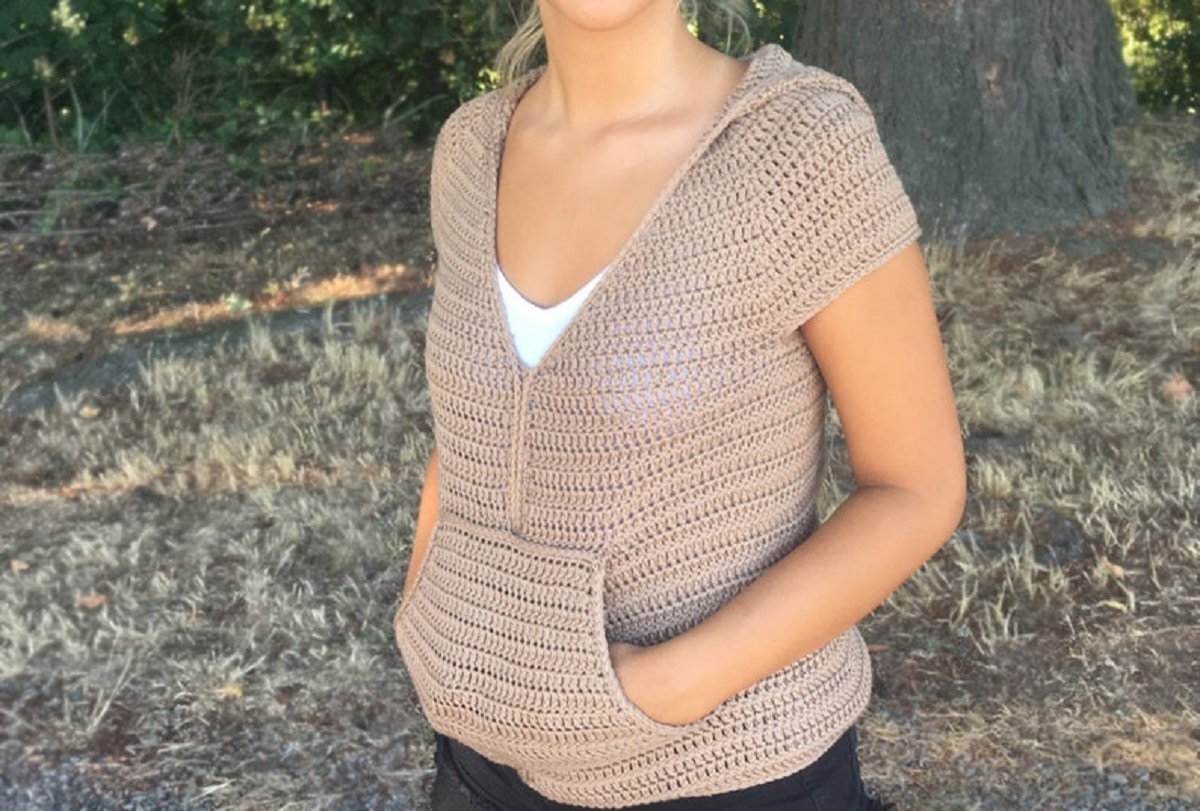 A woman wearing a beige crochet short sleeved hooded sweater with a large pocket on the stomach area.