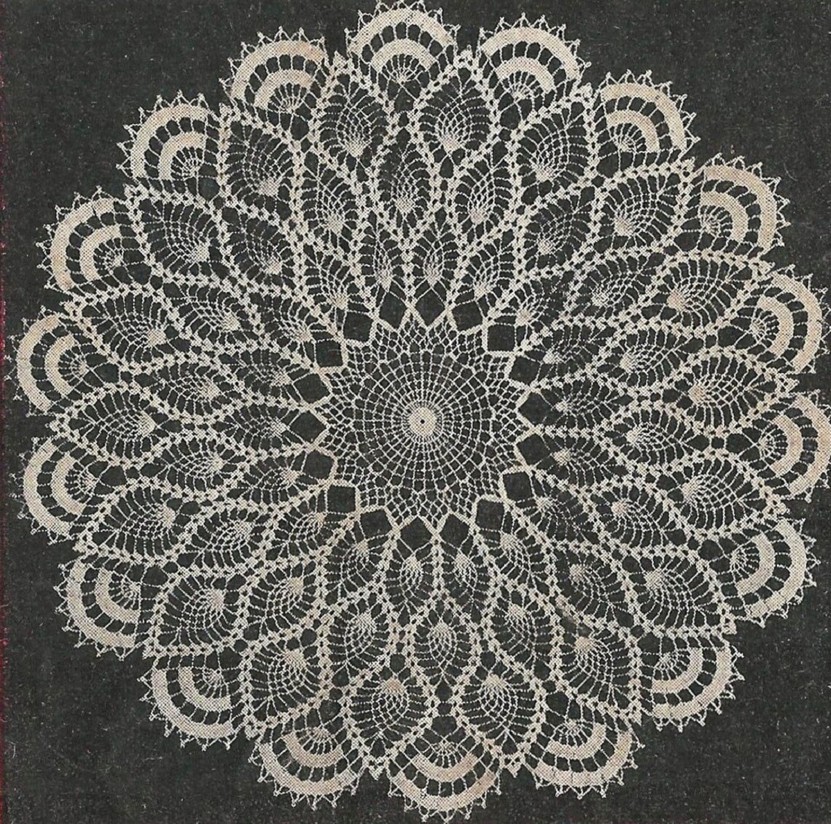 Large crochet doily with a round center and stitched pineapples spreading out in a petal-like design laid on a black background.