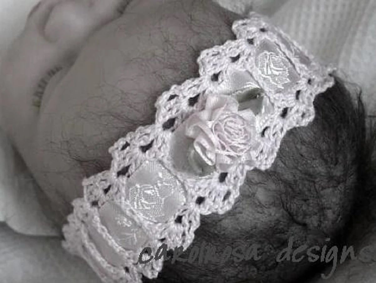  A baby wearing a delicate white crochet headband with lace style detailing on the trim and a crochet flower in the center.