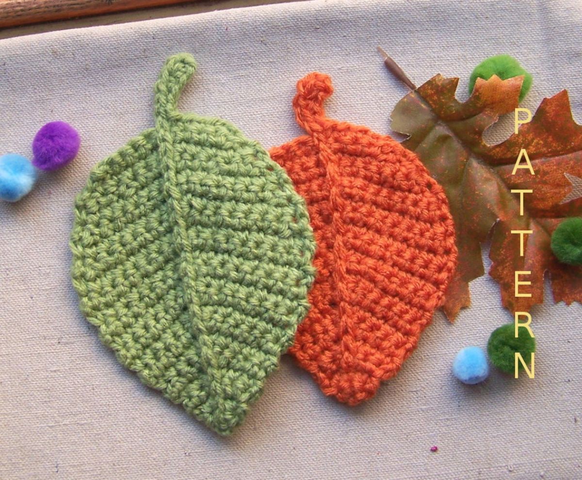Two large crochet autumn leaves in green and orange lying next to each other with small purple and blue balls around them.