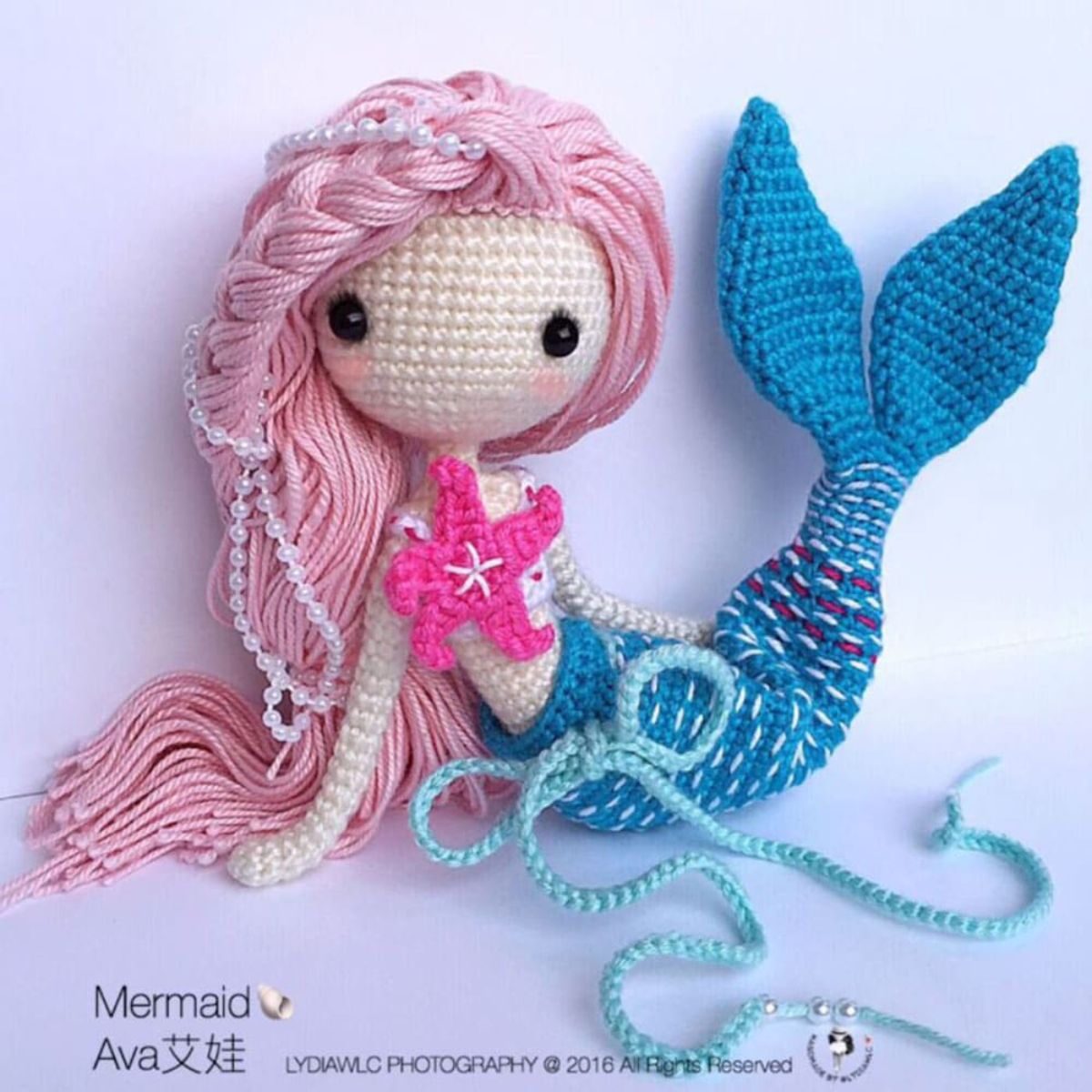Crochet mermaid toy with pale pink hair, a pink starfish, and dark blue crochet tail with a pale blue bow on a lilac background. 