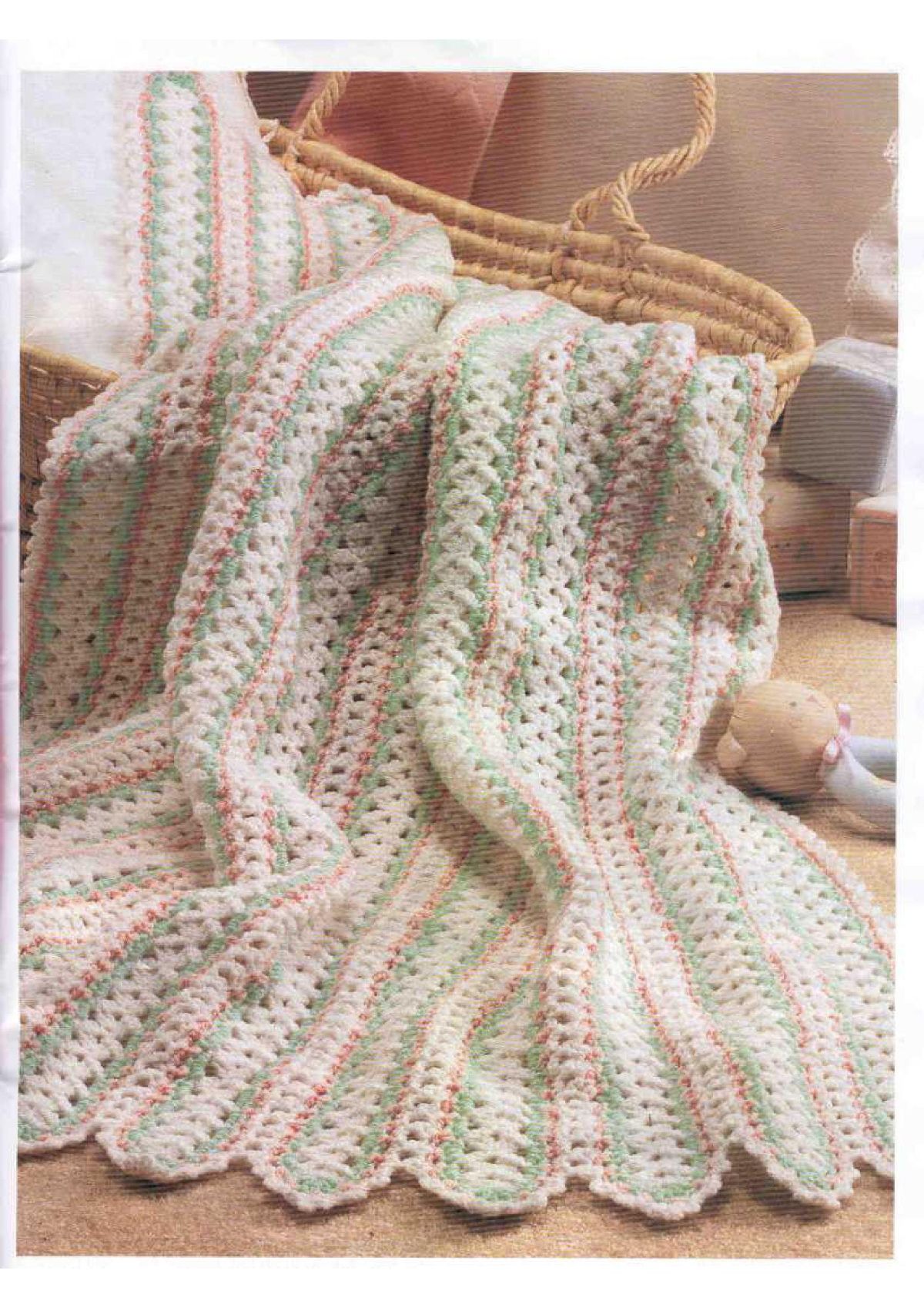 Pale pink, green, and white baby’s afghan with rounded edges spilling out of a brown basket onto the floor. 