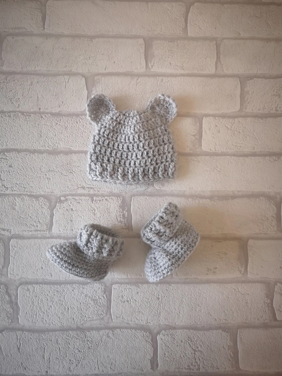  Light gray crochet baby hat with small bear ears at the top of the hat and matching gray booties on a pale brick background.