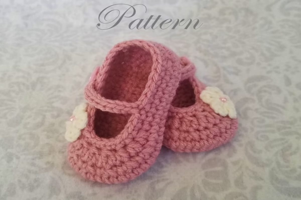  Ballet style crochet pumps for babies in pink with a bar across the middle to secure and a white crochet flower on the side.