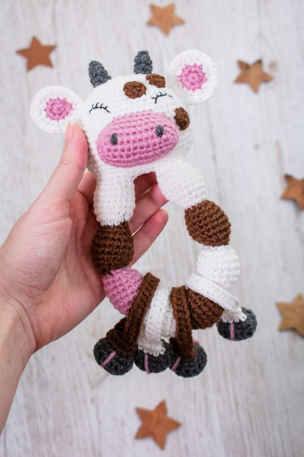 Pink, black, brown, and white crochet cow rattle with a pink nose and gray horns held in a hand with gold stars around it.