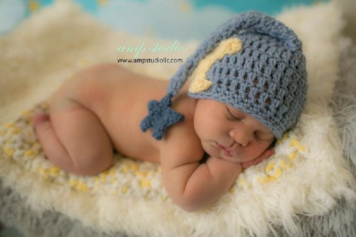  Newborn baby sleeping on a cream rug wearing a blue crochet sleeping cap with a yellow moon on one side and a star dangling on the bottom.