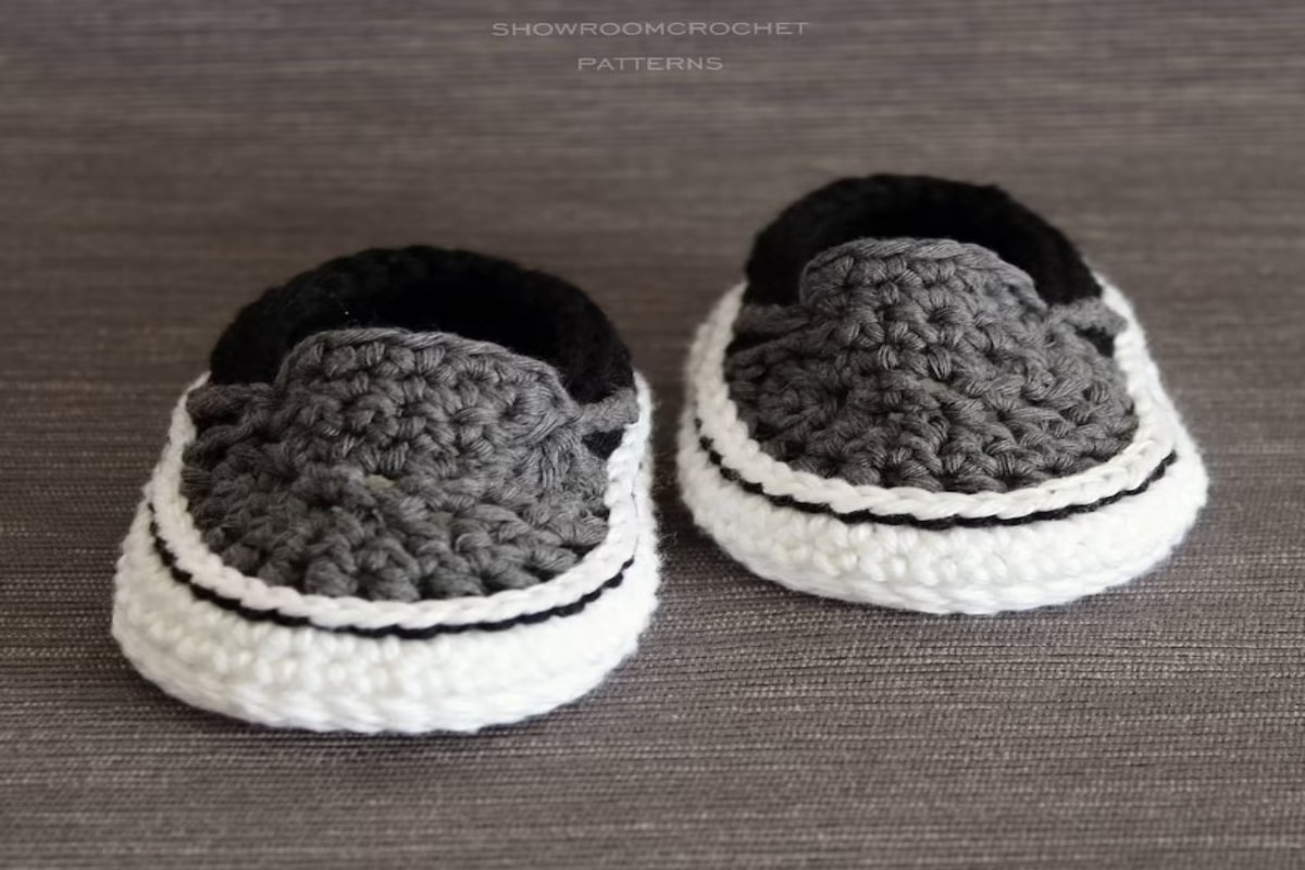 Slip-on crochet baby booties with gray tops, a thick white sole with black trim and black backs on a brown background.