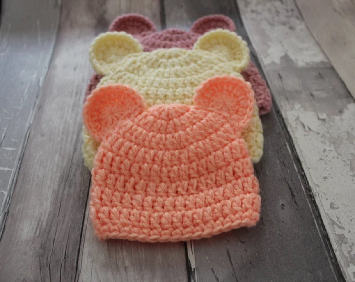 An orange crochet baby hat with small bear ears at the top lying on a pale yellow and pink hat with the same design on a gray wooden background.