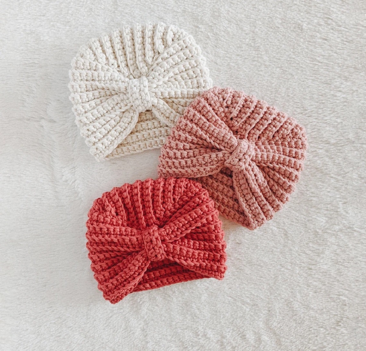 Pink, pale pink, and white turban style crochet baby hats with a bow style in the center on a white soft background.