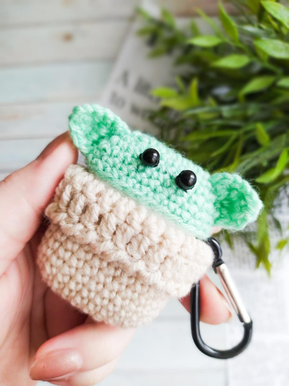 A light green and brown crochet baby Yoda Airpod case held in a hand by some light wooden floor and foliage.