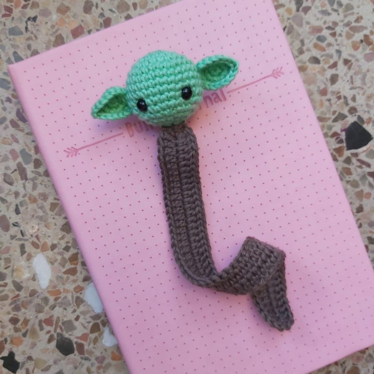 Dark brown crochet bookmark with a small green baby Yoda head at the top lying on a closed pink book.