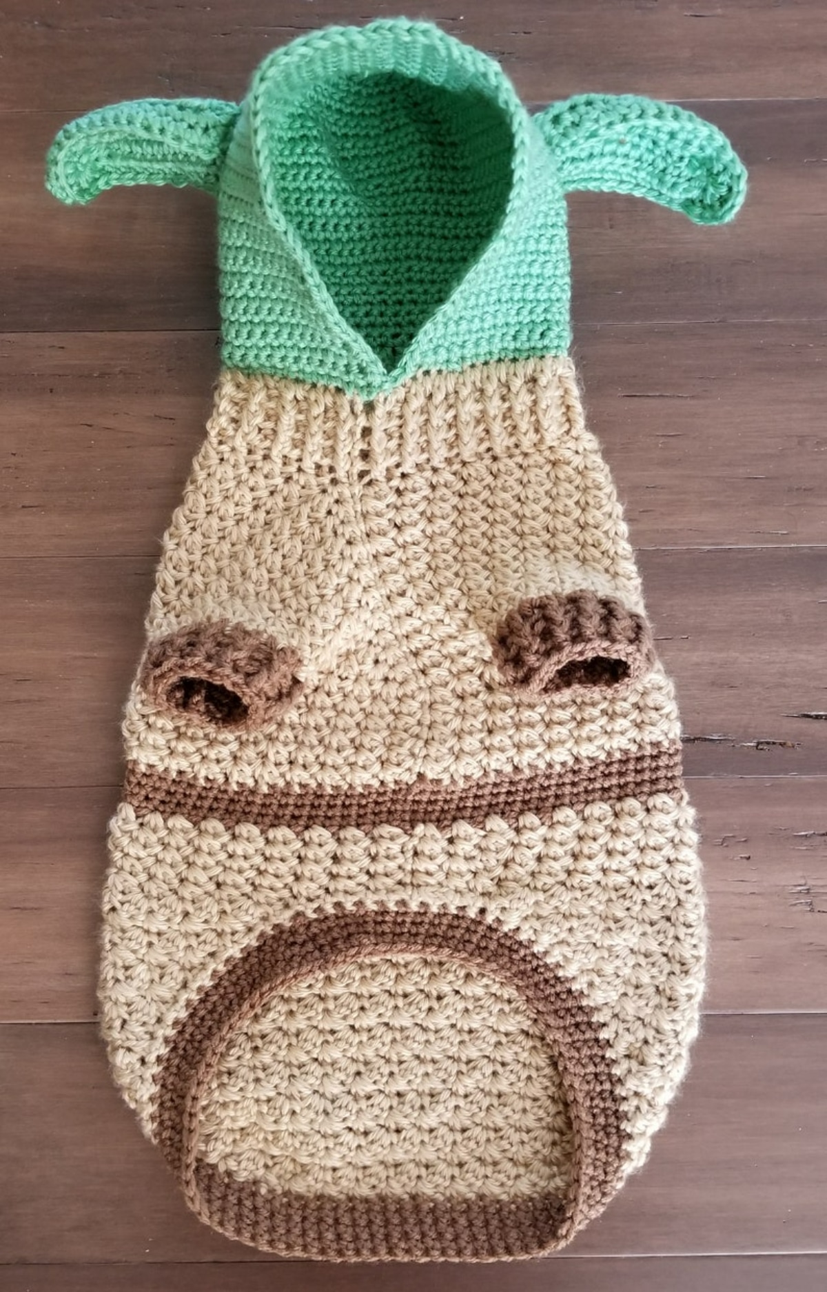A green and brown crochet dog sweater with a green hood with ears, and dark brown cuffs around the legs on a wooden floor.