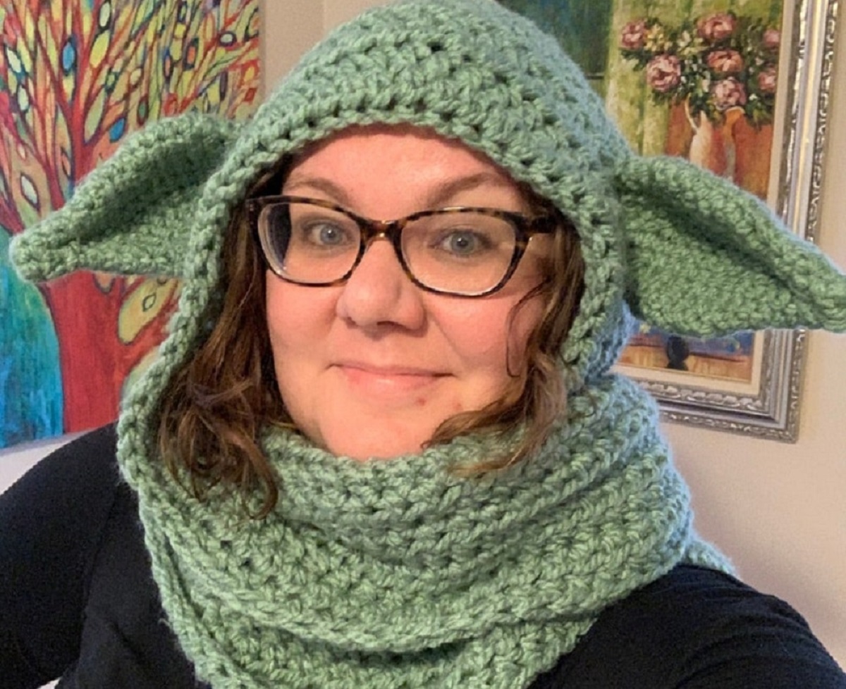 Brunette woman wearing a green crochet baby Yoda hooded scarf with large ears on either side.