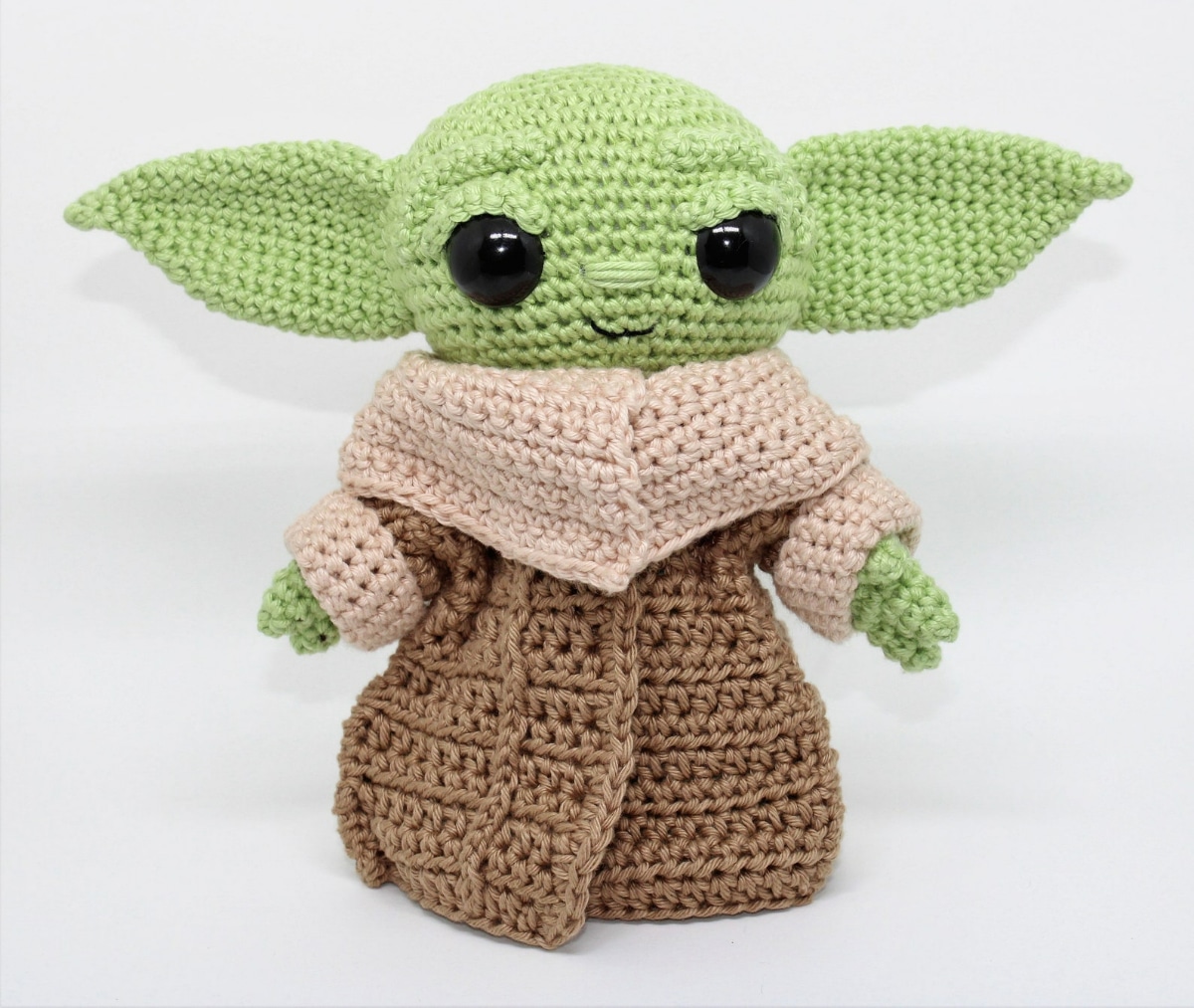 Small green crochet baby Yoda wearing a brown robe with a pale pink collar and cuffs standing on a white background.