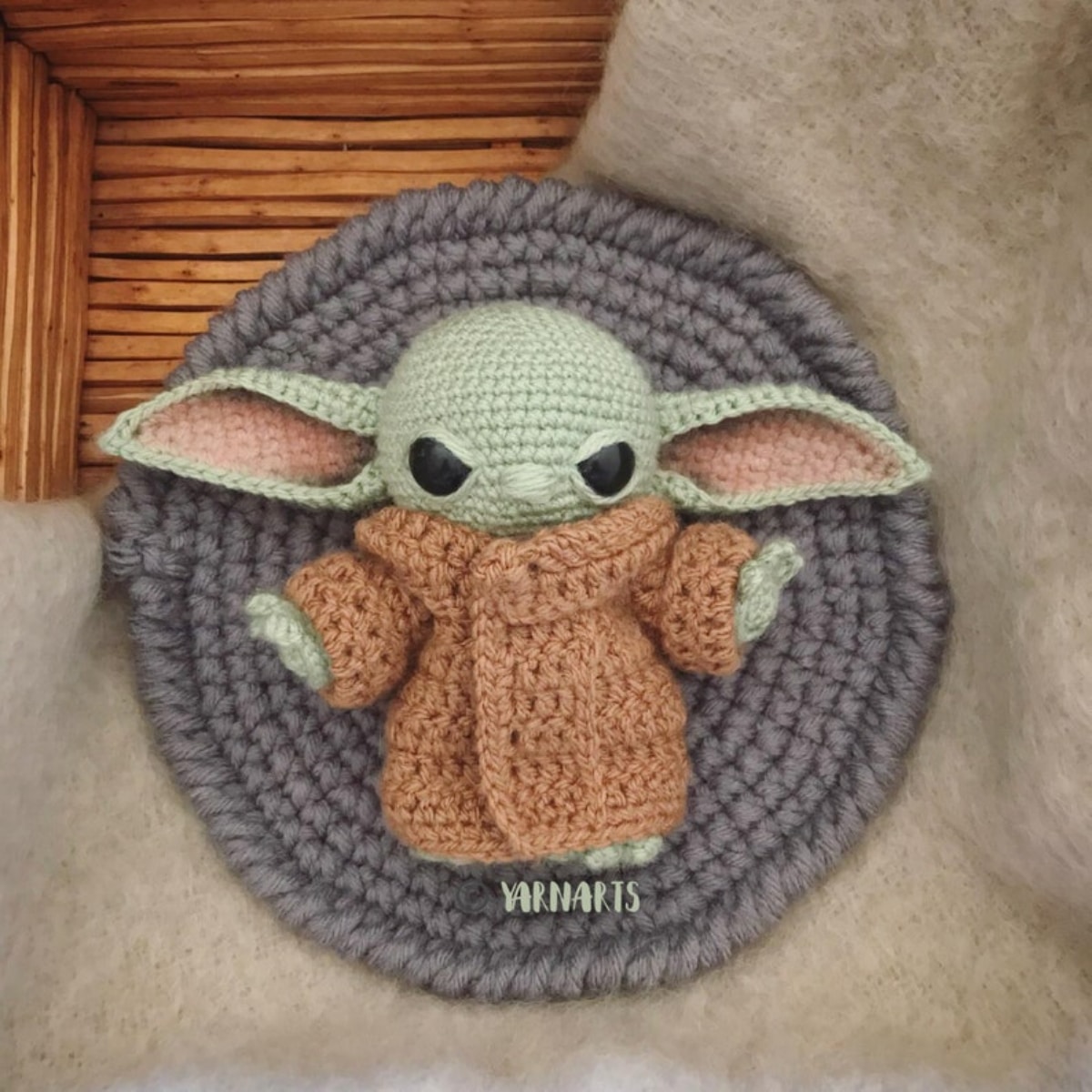Small green crochet baby Yoda with pink ears wearing a brown robe and lying in a gray crochet pod.