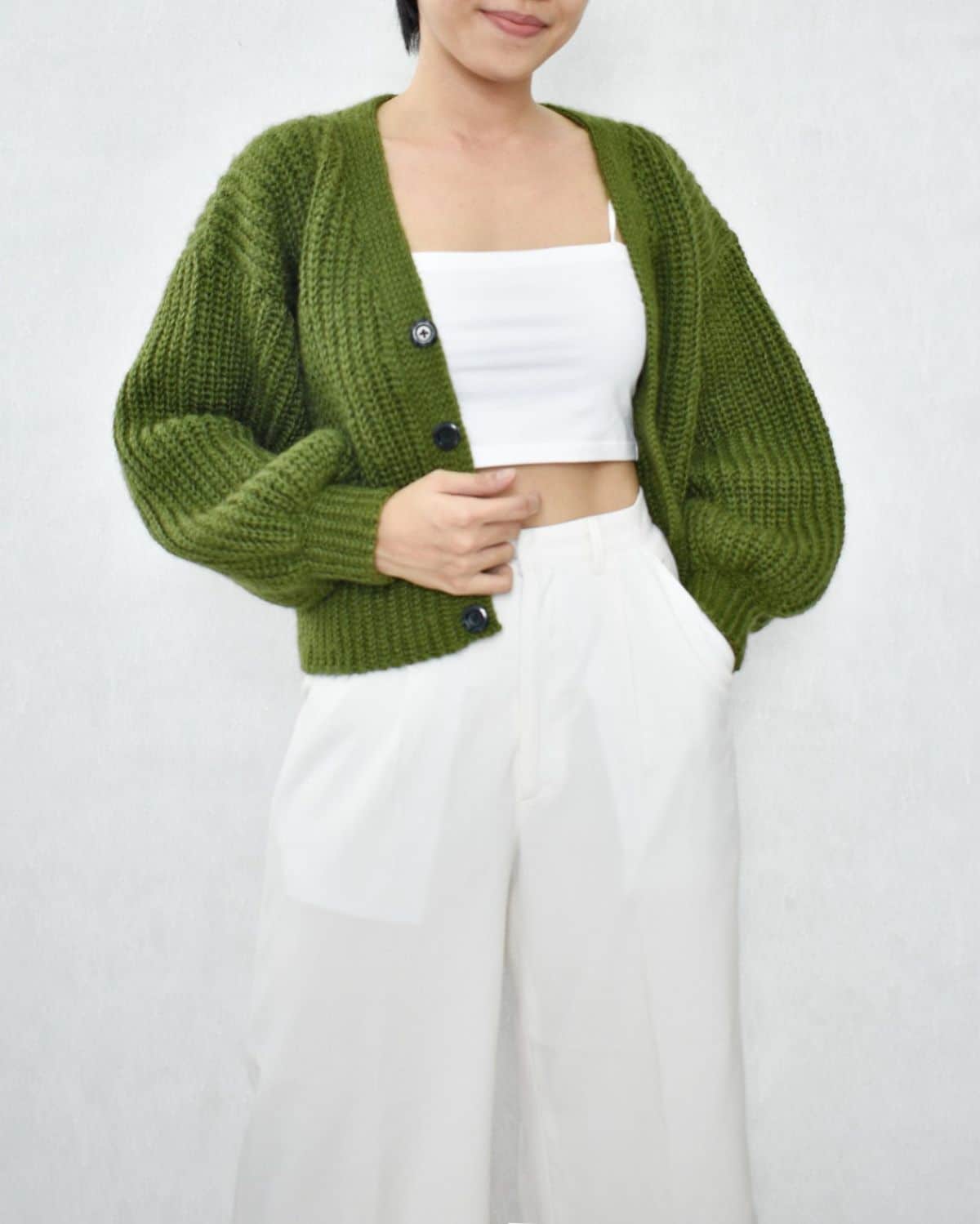 A woman in a white top and trousers wearing a chunky crochet green hip-length cardigan with balloon sleeves and cuffs at the bottom.